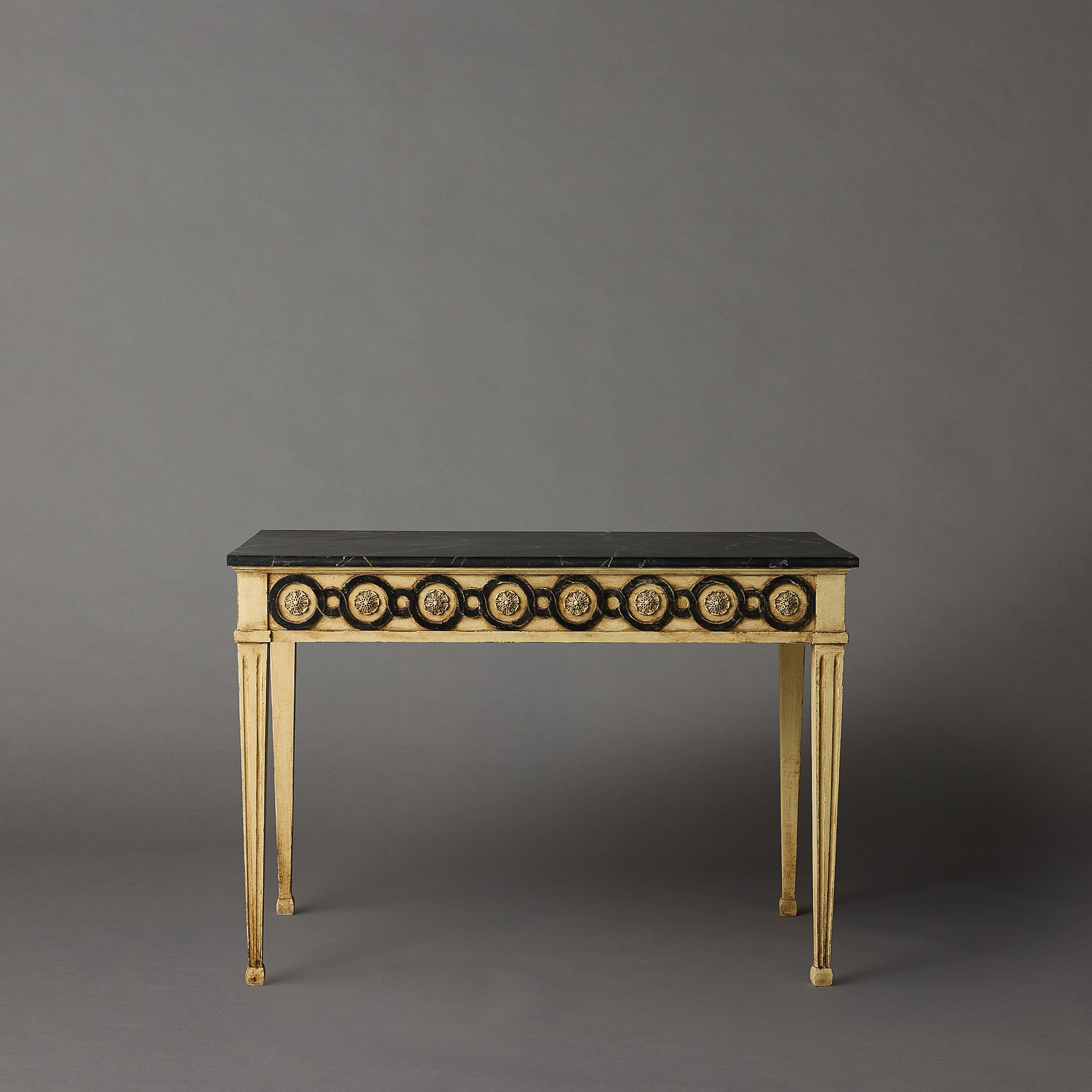 A classic French console table with a black marble top, gold-painted frame, and circular black and gold inlay details on the front apron, set against a solid gray studio background.
