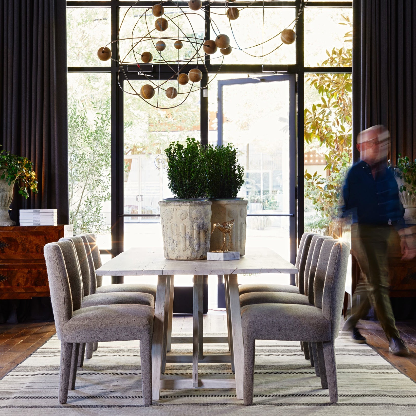 A dining room scene with a rectangular light wood table, four upholstered gray chairs, a large chandelier, and a blurred person walking in the background near tall windows and dark curtains.