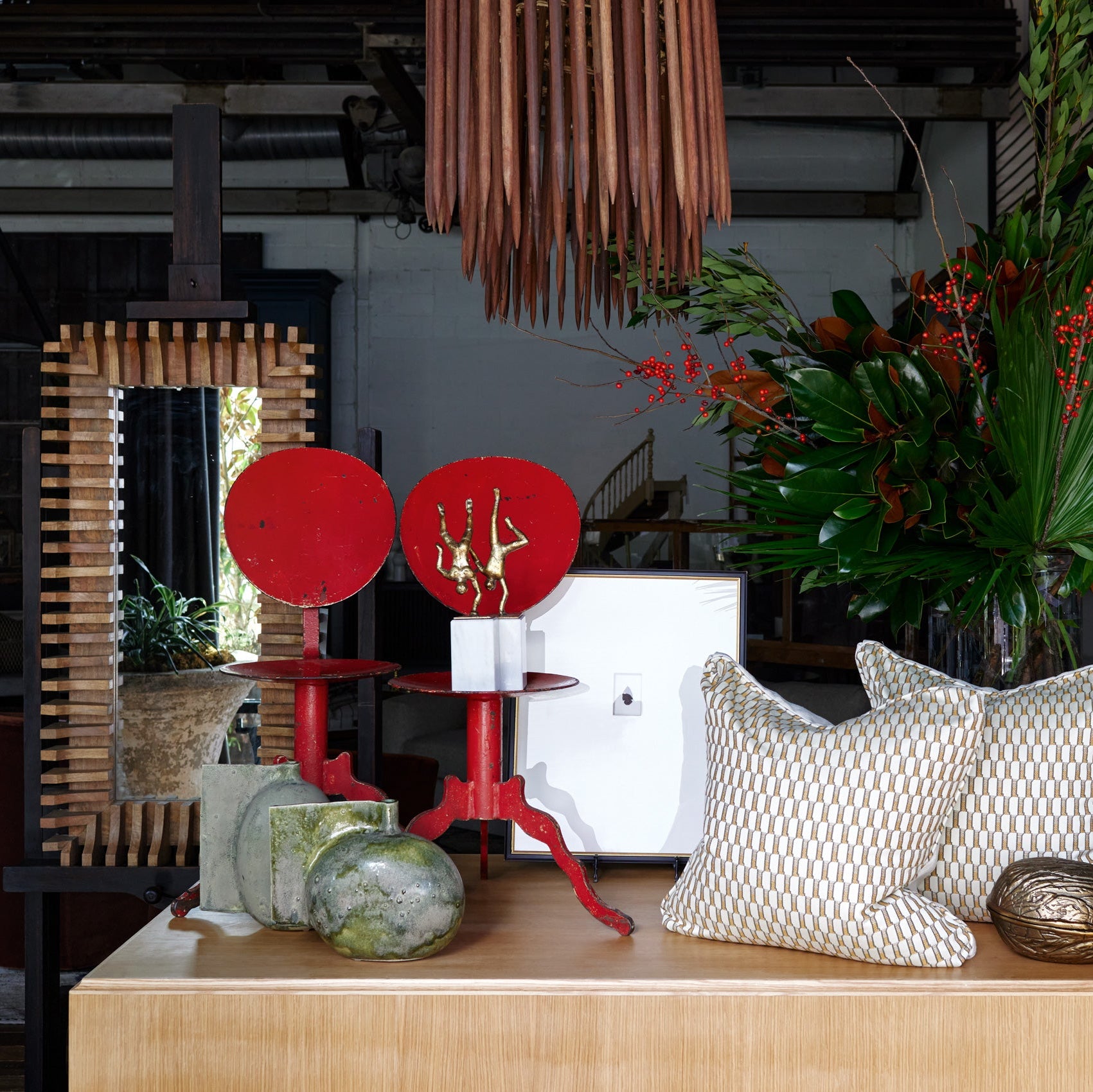 A styled interior scene showing a light wood console table with two cream patterned throw pillows, a red decorative object, and a large floral arrangement in the background.