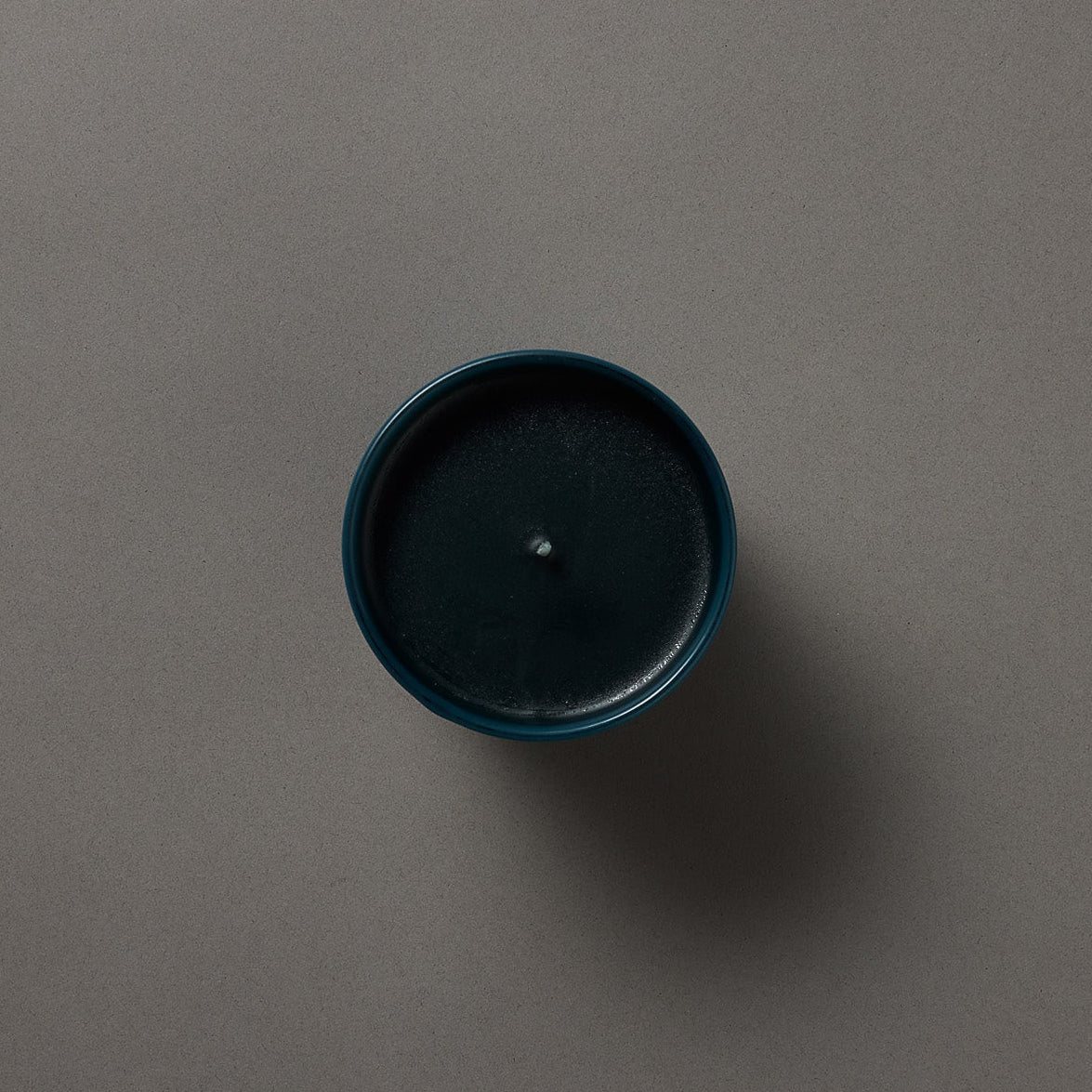 A top-down view of a dark teal blue candle in a matching round container, centered on a solid dark gray background.