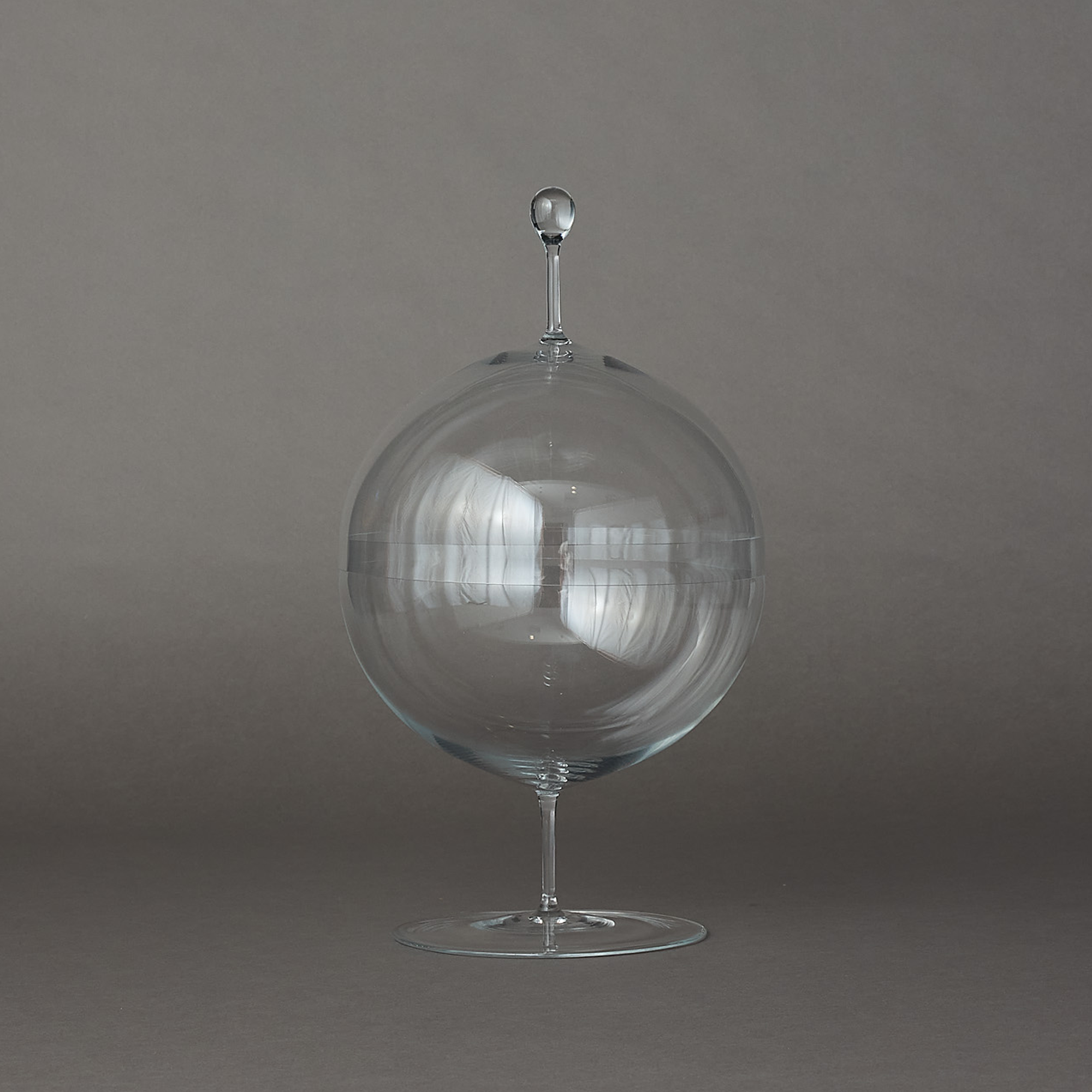 A clear glass decorative object with a large spherical body, a narrow stem, and a small round finial on top, photographed against a plain gray studio background.