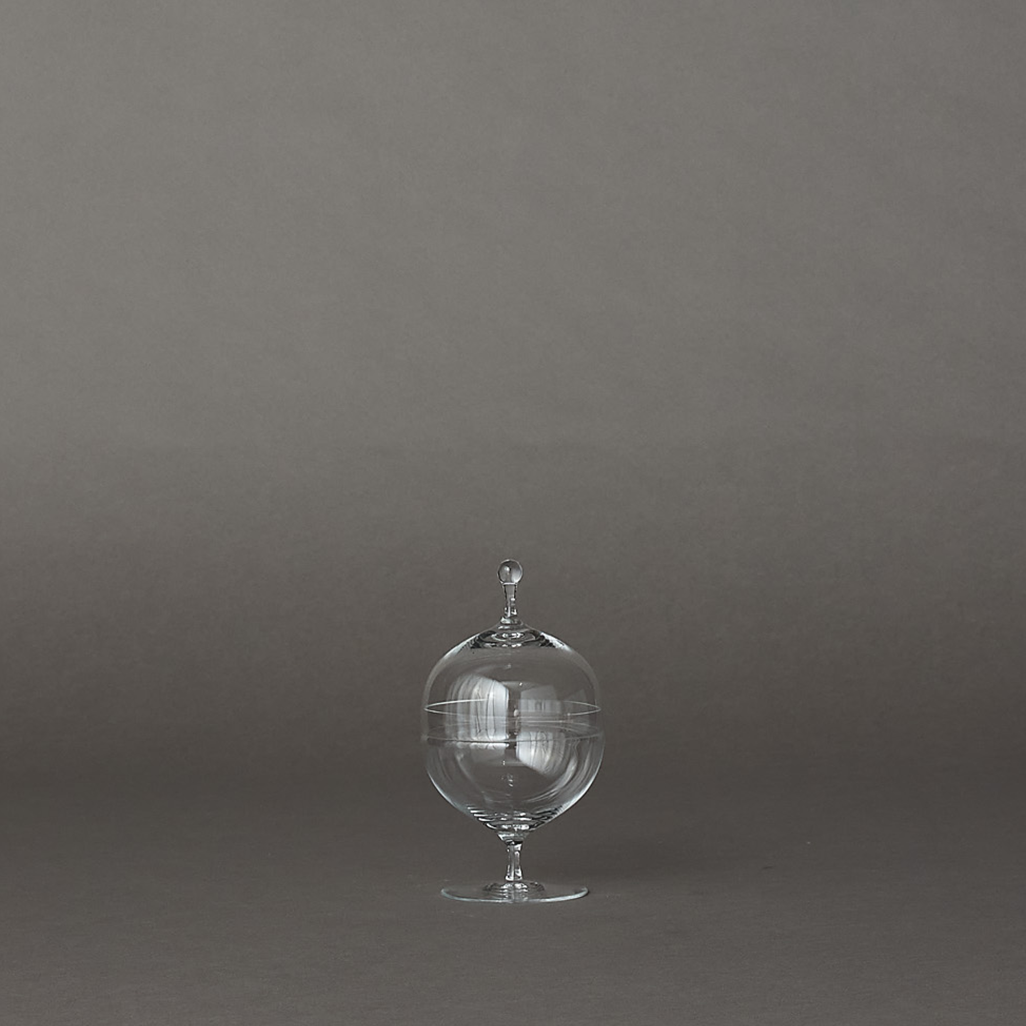 A clear glass decorative container with a rounded body, a small pedestal base, and a matching screw-top lid, photographed against a smooth dark gray studio background.