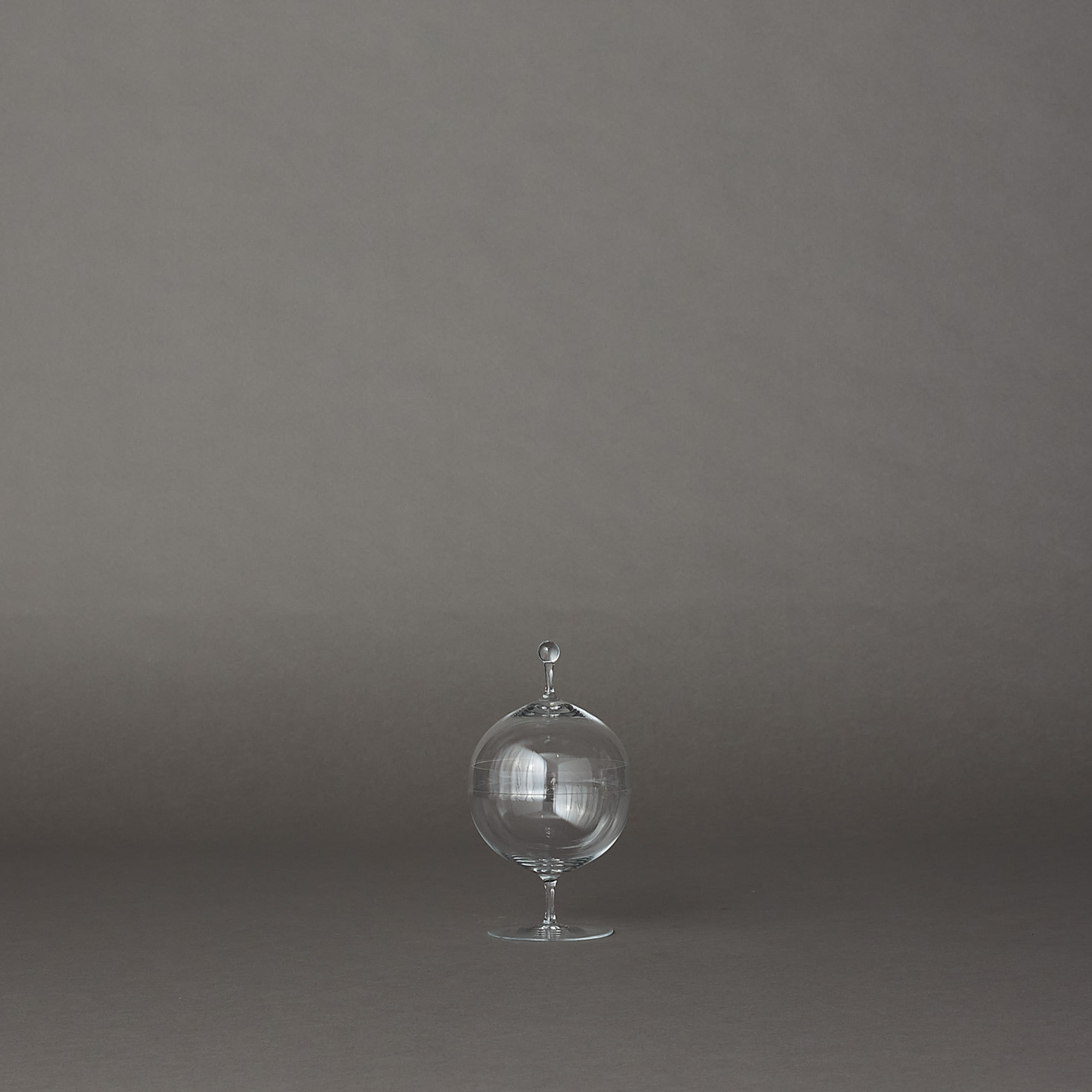 A clear glass decorative jar with a lid and pedestal base, photographed against a plain gray studio background.