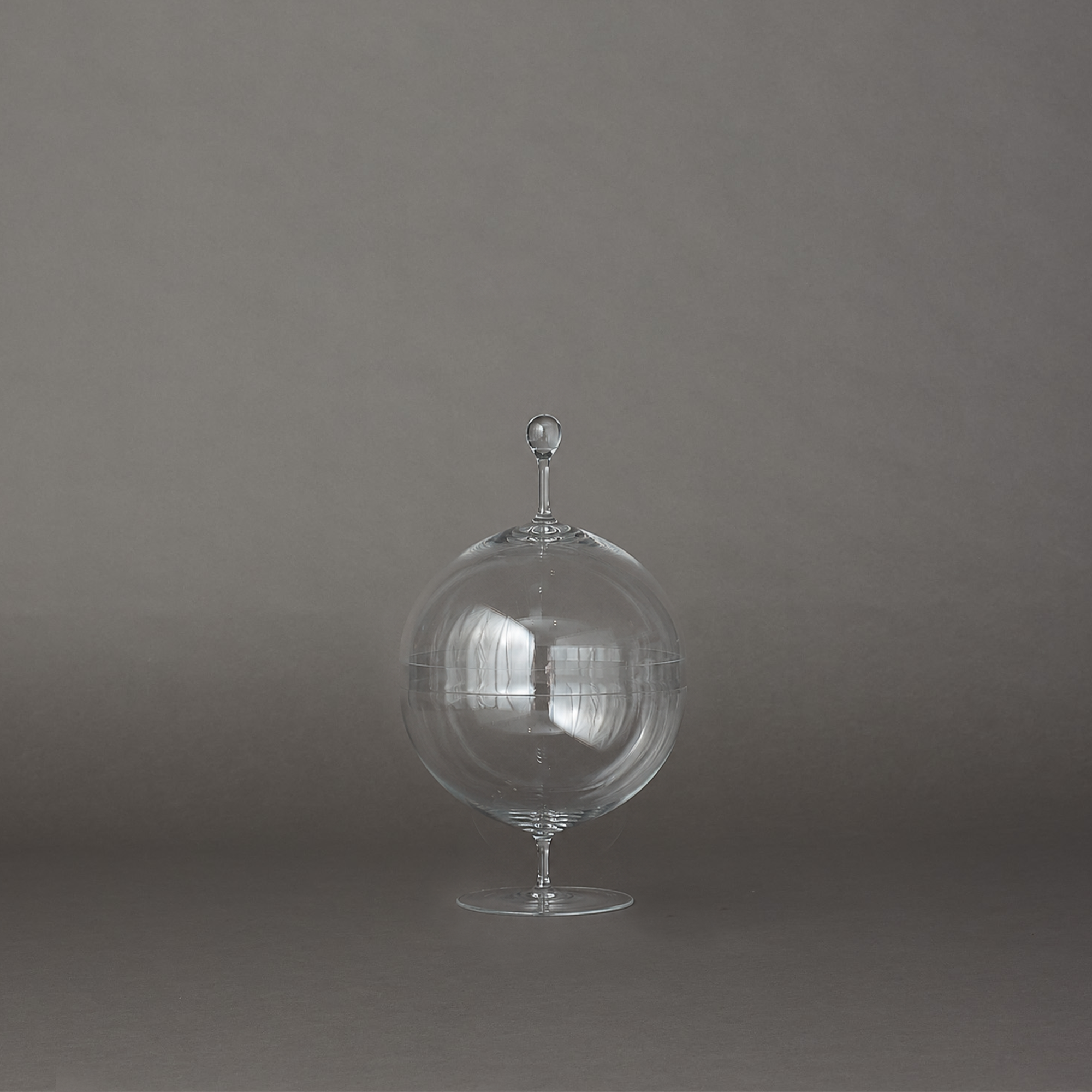 A clear glass decorative object with a rounded bulbous body, narrow stem, and small finial on top, photographed against a smooth gray studio background.
