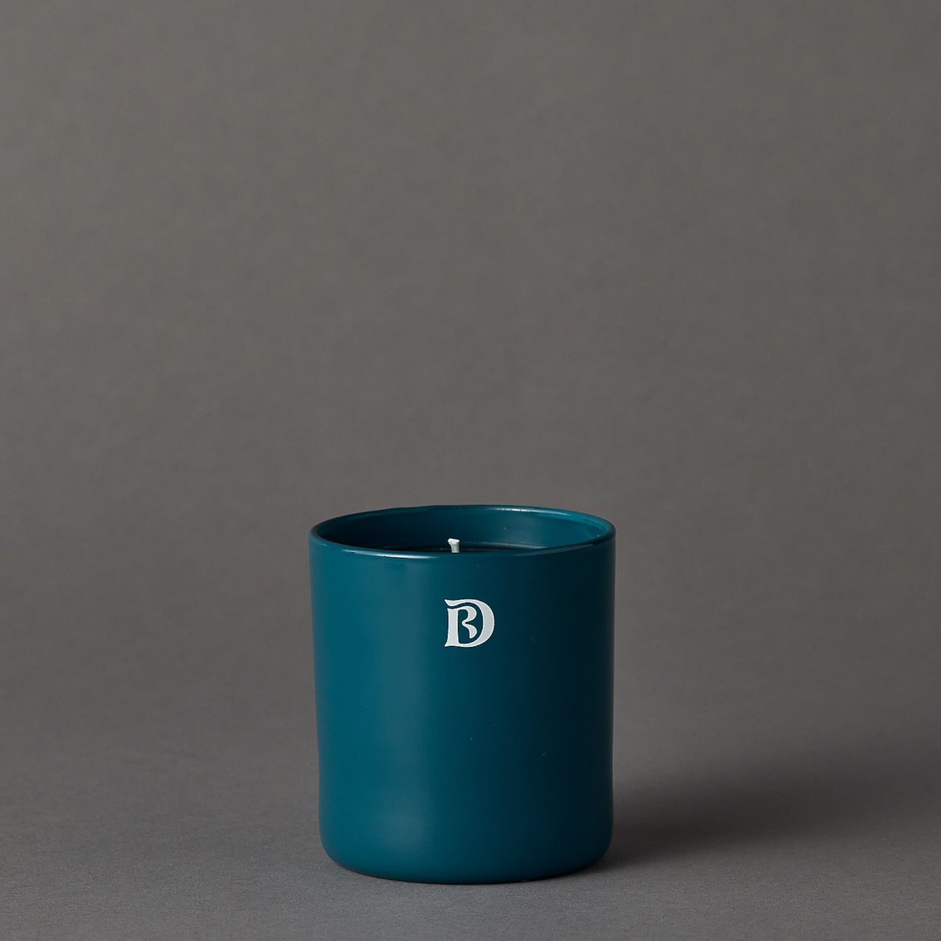Teal candle in a cylindrical container with a logo on a gray background