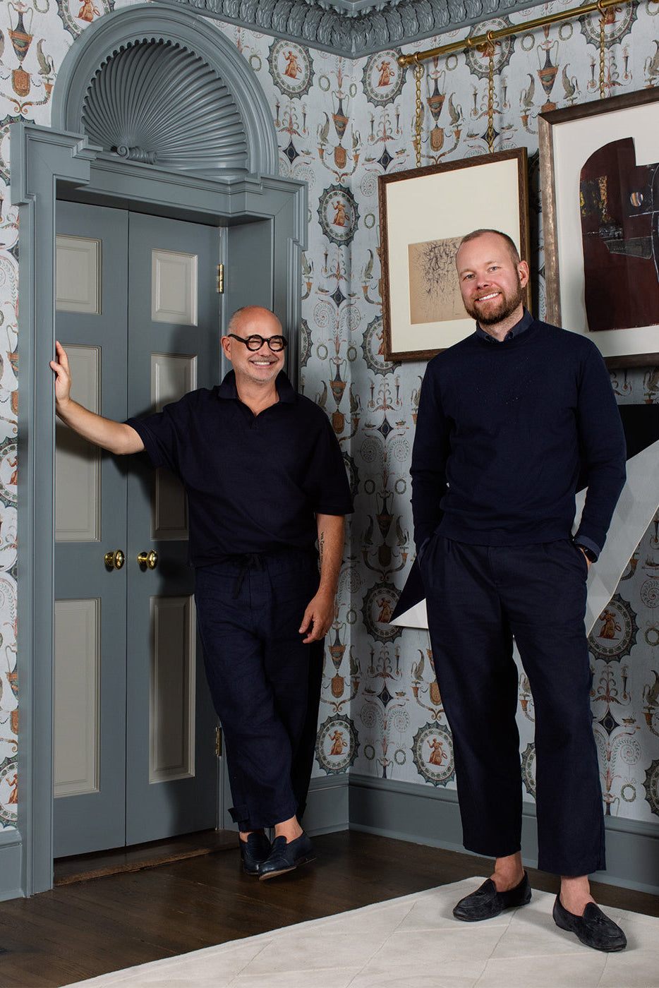 Two men standing in a room with decorative wallpaper and framed pictures.